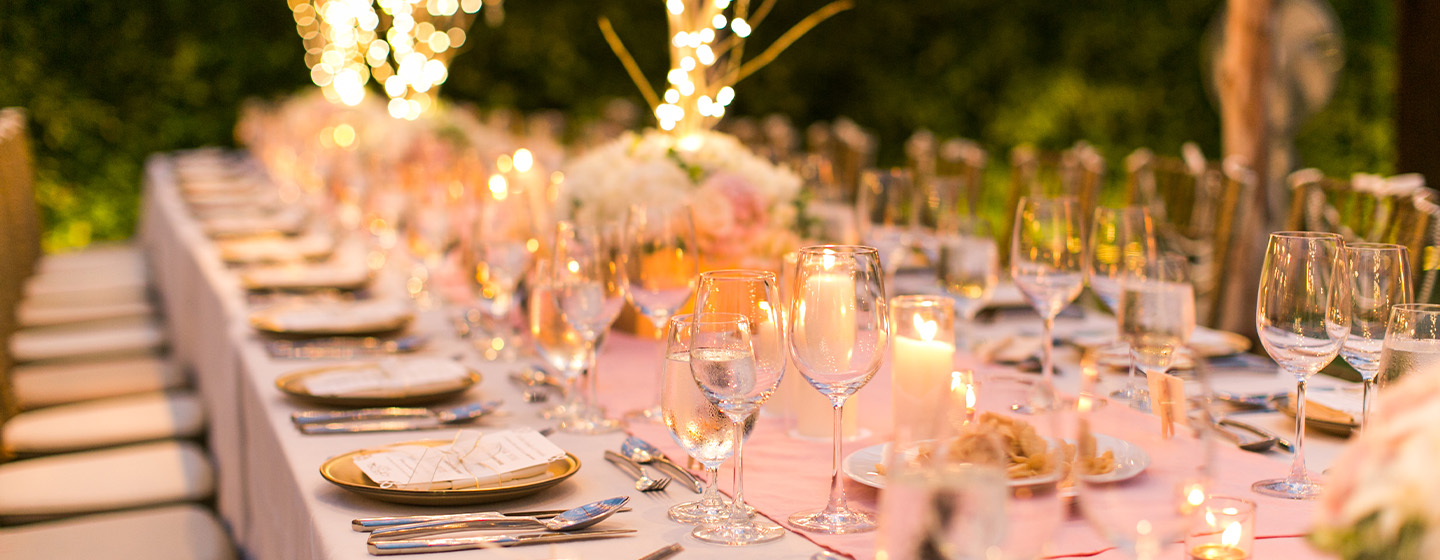 Elegant outdoor dining setup with a long table adorned with pink flowers, candles, and glassware, creating a warm, romantic atmosphere. Soft lighting enhances the mood.