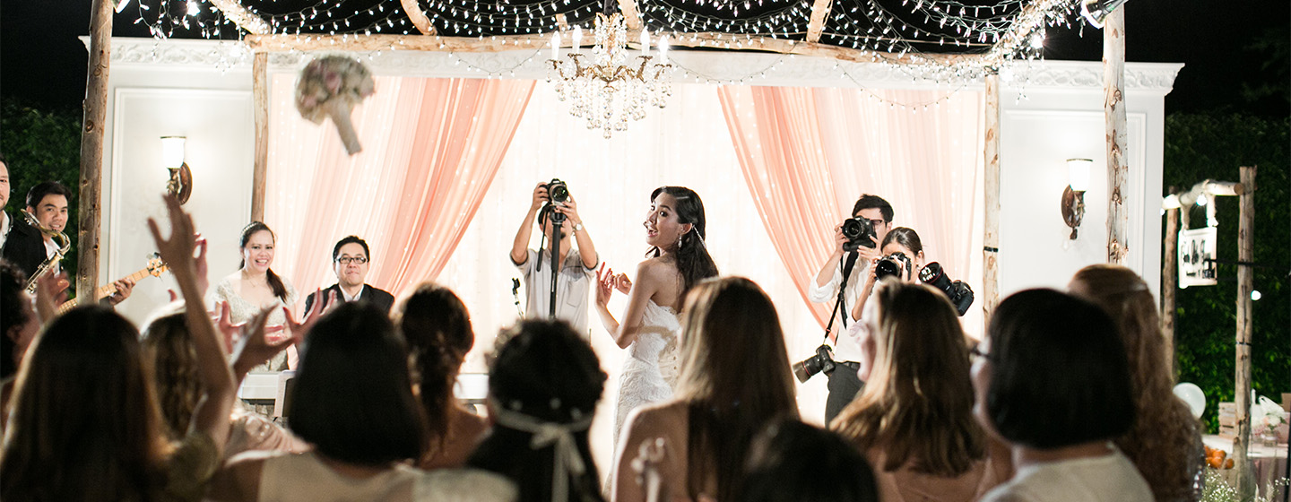 Bride in wedding dress tossing bouquet to a group of excited guests. Photographers capture the moment against a backdrop of pink drapes and chandelier.