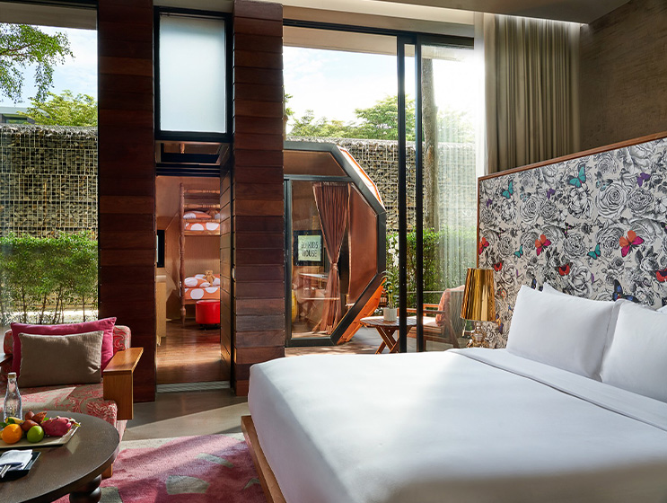 Modern hotel room with a large bed, patterned butterfly wallpaper, and a cozy seating area. Glass doors lead to a lush outdoor patio, creating a serene ambiance.