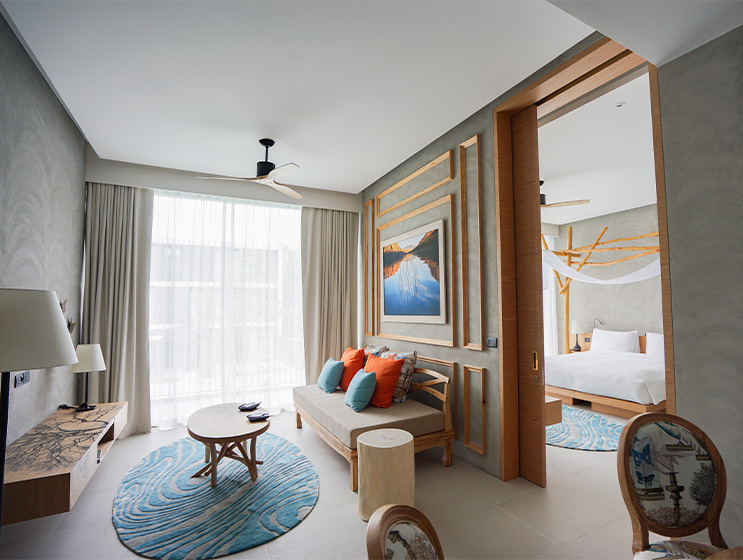 Modern hotel room with natural light, featuring a sofa with orange and teal cushions, a round table, and a bed with white linens. Cozy and inviting.