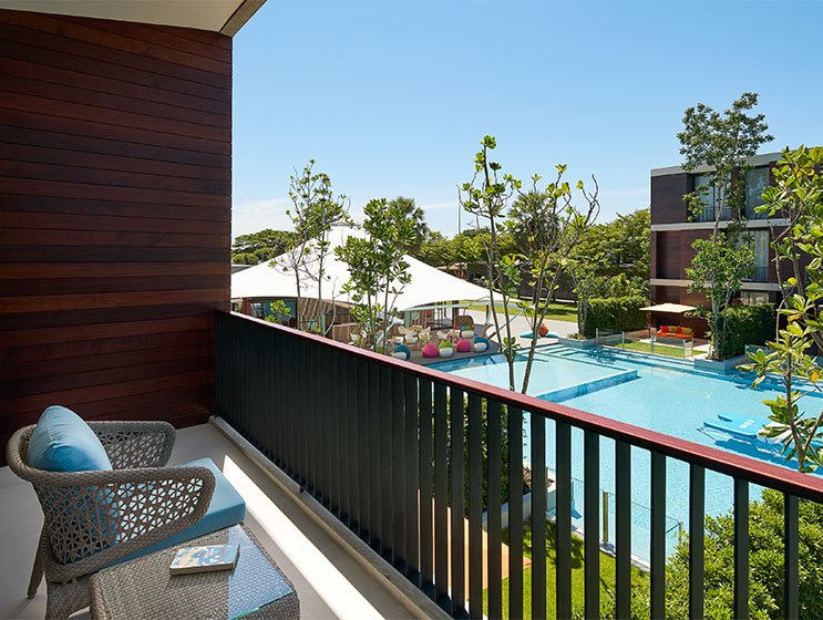 View from a balcony overlooking a sunny pool area with lounge chairs, umbrellas, and surrounding greenery. A peaceful, relaxing atmosphere is conveyed.