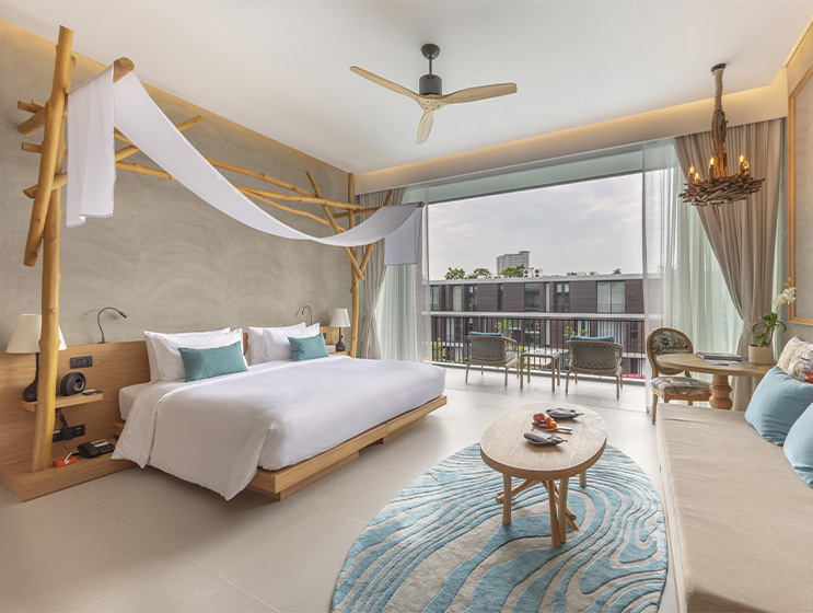 A spacious, serene bedroom with a large bed, light wood furniture, and a blue rug. Sunlight filters through sheer curtains, creating a calm, airy atmosphere.