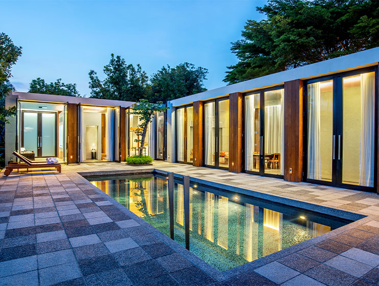Modern house with large glass doors overlooking a tranquil pool at dusk. The warm indoor lighting contrasts with the cool blue exterior, creating a serene ambiance.
