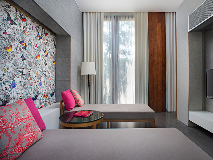Modern room with two gray daybeds featuring pink cushions. A floral and butterfly mural adorns one wall. A sheer-curtained window and tall lamp add light.
