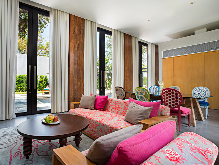 Bright, modern living room with pink-patterned sofas, wooden coffee table, and tall windows with white curtains. Light and airy ambiance.