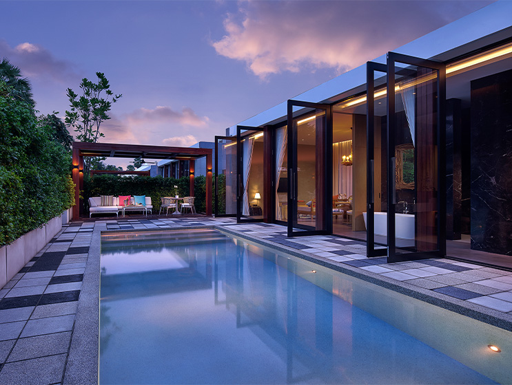 Luxurious poolside view at dusk, with open glass doors revealing an elegant interior. The serene pool reflects a vibrant, cloudy sky, evoking tranquility.