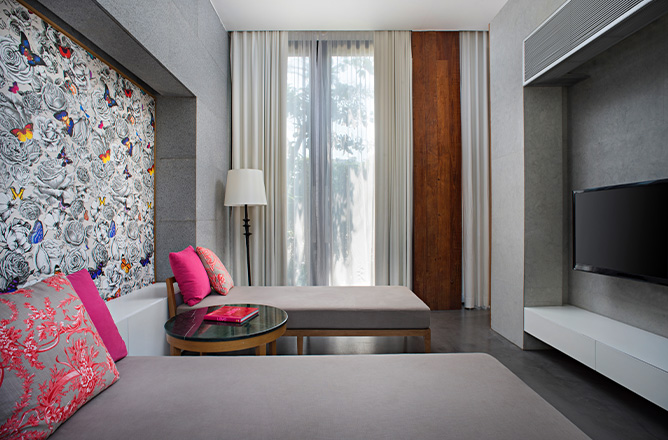 http://Modern%20room%20with%20two%20gray%20daybeds%20featuring%20pink%20cushions.%20A%20floral%20and%20butterfly%20mural%20adorns%20one%20wall.%20A%20sheer-curtained%20window%20and%20tall%20lamp%20add%20light.