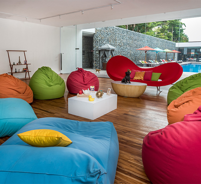 Colorful bean bags and a wavy red couch in a cozy, modern lounge with wooden floors. A small table holds fruit, and there's a view of a pool outside.