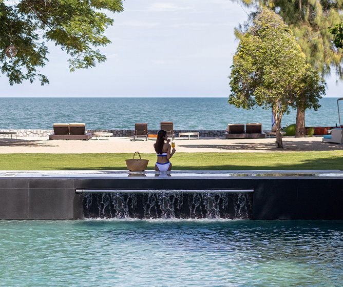 A woman stands by a sleek infinity pool with a flowing waterfall, overlooking an ocean view. Trees frame the scene, evoking a tranquil, luxurious ambiance.