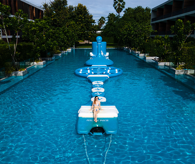A woman lounges on a white inflatable in a long blue pool with a large blue Buddha statue at the end. Lush trees line the poolside. Relaxing vibe.