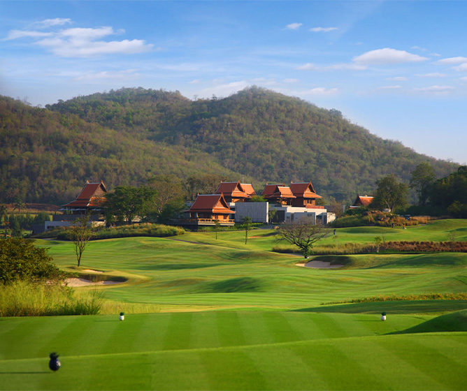 A lush, green golf course under a clear blue sky, with rolling hills and red-roofed buildings in the background, conveying a serene and peaceful atmosphere.