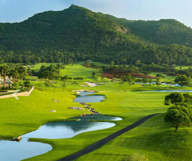 Lush green golf course with small ponds and winding paths, set against a backdrop of forested hills under a clear blue sky, evoking tranquility.