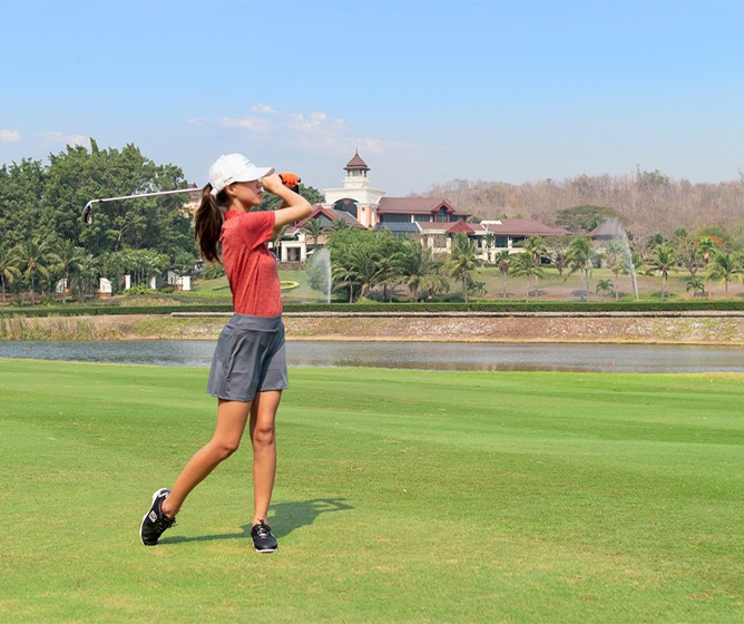 A golfer in a red shirt and gray shorts follows through on a swing on a lush course, with trees and a clubhouse in the background under a clear sky.