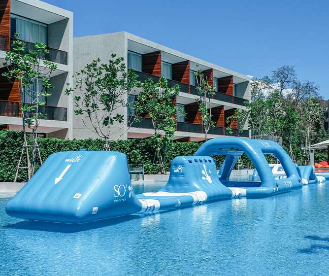 Large inflatable water obstacle course floats in a clear blue pool. Modern apartment building and lush greenery form the backdrop under a sunny sky.