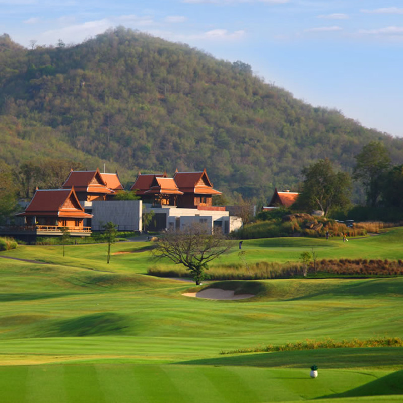 Scenic view of a lush green golf course with rolling hills, traditional-style buildings with red roofs, and wooded hills in the background under a blue sky.