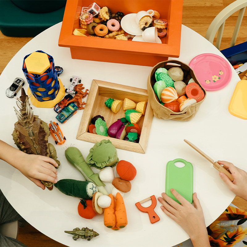 A round table with children's hands playing with toy vegetables and kitchen tools. A colorful mix of play food and utensils creates a fun, playful atmosphere.