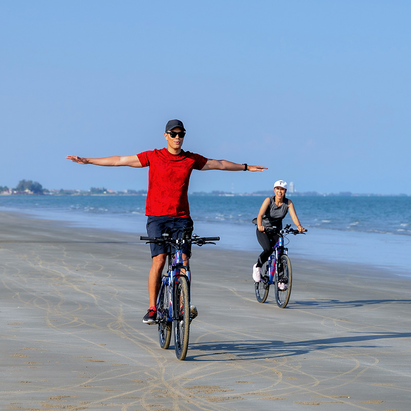 Two people ride bikes on a sandy beach. The person in front extends arms for balance, conveying freedom and joy under a clear blue sky.