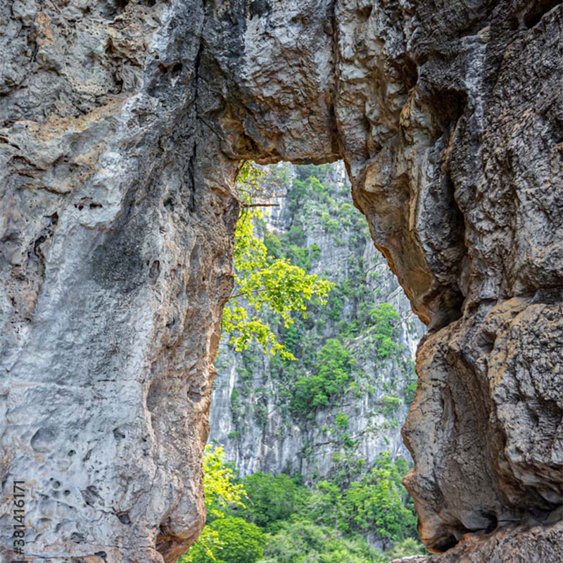 A natural rock arch frames a view of lush green foliage and distant cliffs. The scene evokes a sense of tranquility and natural beauty.