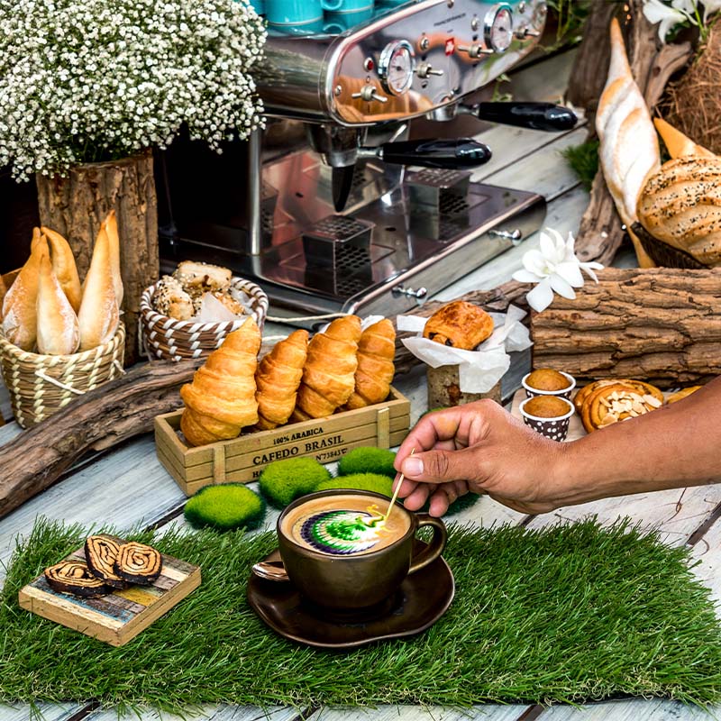 A hand stirs coffee with latte art on a grassy surface. Nearby are pastries, bread, flowers, and a coffee machine, conveying a cozy, rustic vibe.