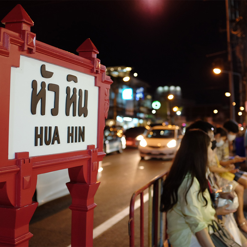 Night scene in Hua Hin with a red sign in the foreground. People sit and stand near a busy street lined with lights, creating a lively, bustling atmosphere.