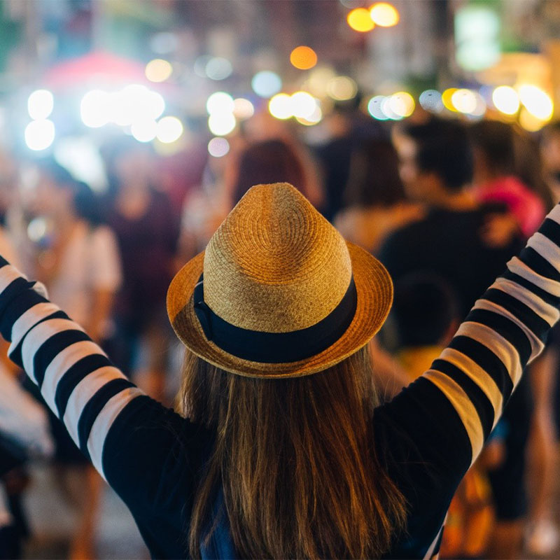 A person in a yellow hat and striped shirt raises their arms in a bustling, brightly lit night market, conveying excitement and energy.