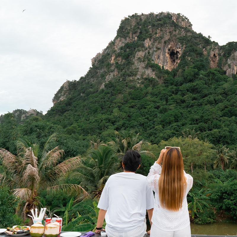 A couple in white clothing stands at a picnic table, admiring a lush green mountain with a cave, under a cloudy sky. The scene is peaceful and serene.