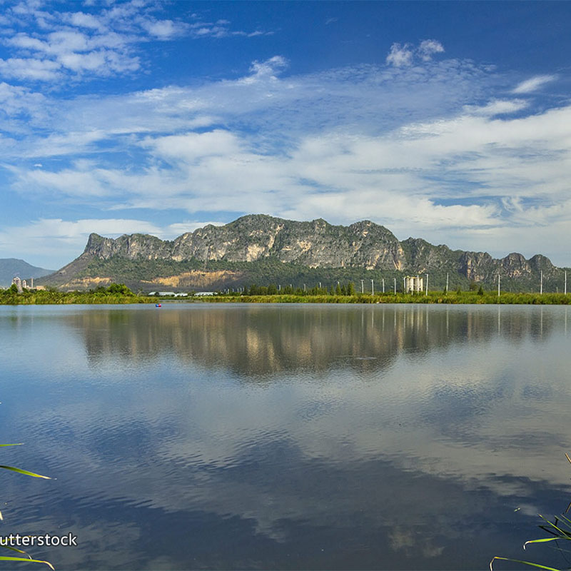 A serene landscape showing a mountain range under a blue sky with scattered clouds, reflected on the calm water of a lake in the foreground.