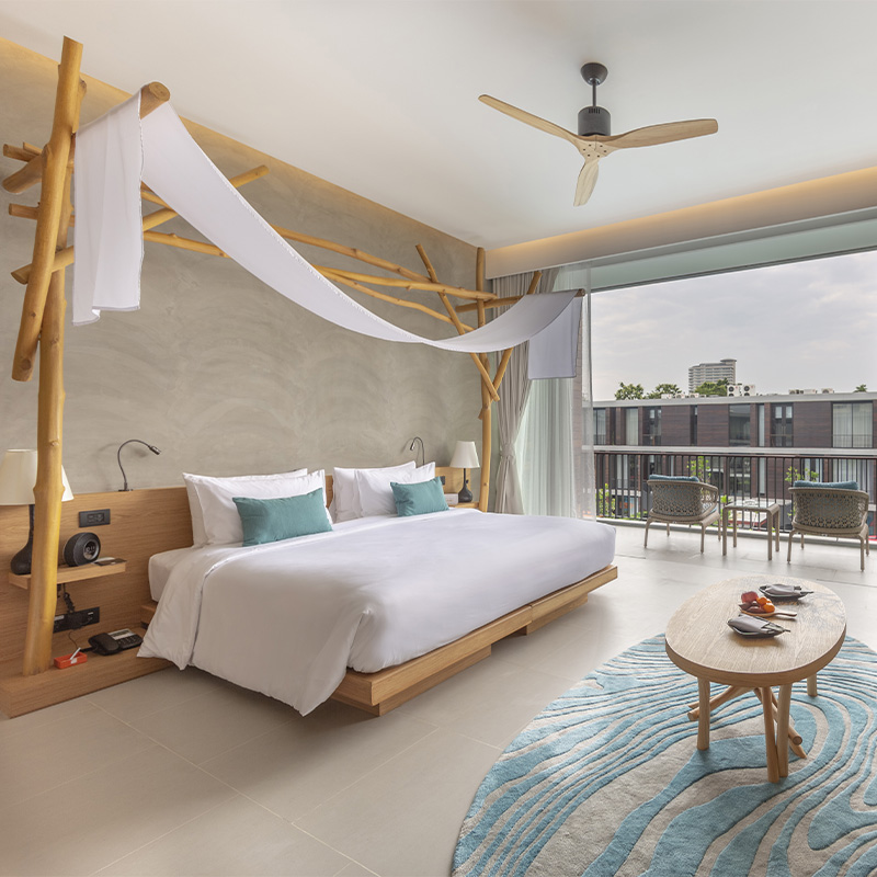 Modern, airy hotel room with a white bed adorned with teal pillows. Bamboo decor frames the headboard. A glass door leads to a balcony with chairs and a city view. The room features a ceiling fan and a light wood coffee table with fruit, evoking a calm, inviting atmosphere.