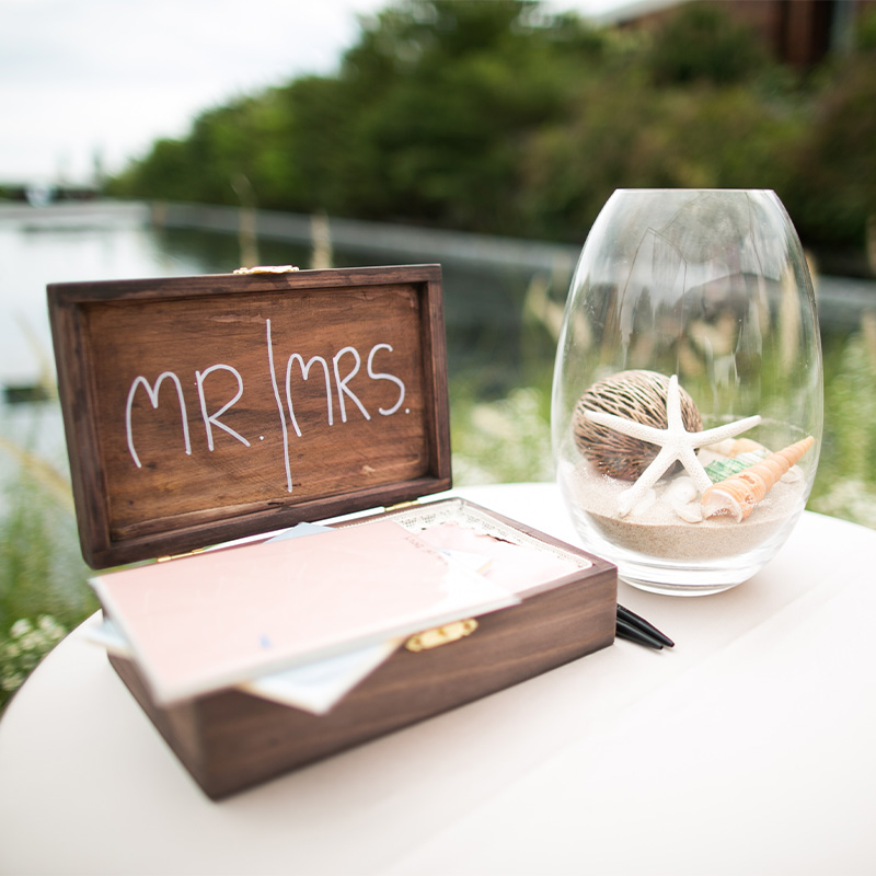 Wooden box labeled "Mr & Mrs" open on table beside large glass vase with sand and shells. Background shows a blurred, lush riverside setting.