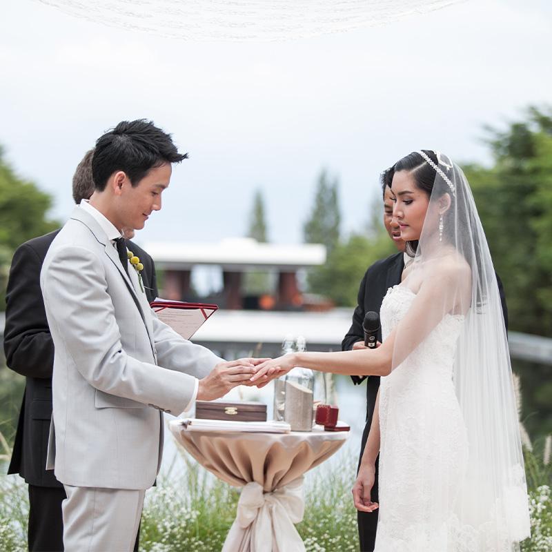 A bride and groom exchange rings during an outdoor wedding ceremony. The groom wears a light gray suit, and the bride is in a white dress with a veil, appearing emotional.