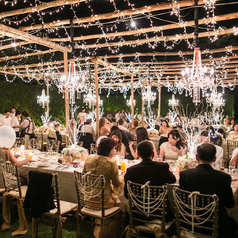 Elegant outdoor banquet with guests seated at long tables under twinkling lights and chandeliers. The setting exudes a festive and joyful atmosphere.