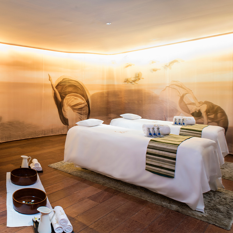 Calm spa room with two massage tables draped in white sheets and striped runners, surrounded by a serene mural of figures and birds. Soft lighting enhances the tranquil atmosphere.