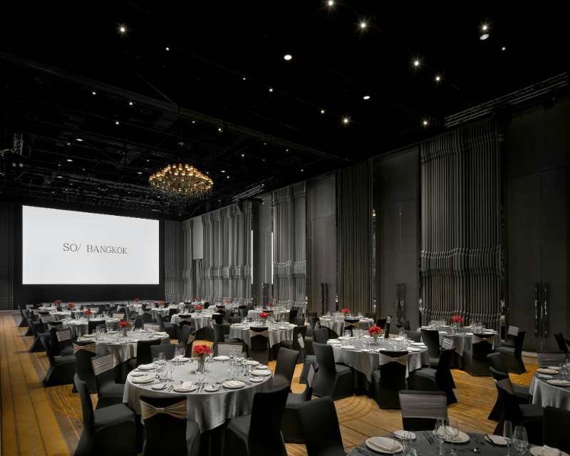 http://Several%20round%20fully%20set%20tables%20sit%20under%20a%20chandelier%20in%20a%20large%20ballroom%20with%20a%20large%20screen%20hung%20at%20the%20front.
