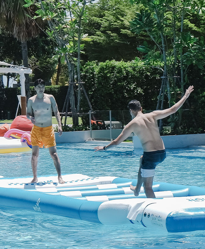 Two people enjoying a pool on inflatable logs. One steadies himself while the other walks, surrounded by lush greenery and a sunny atmosphere.