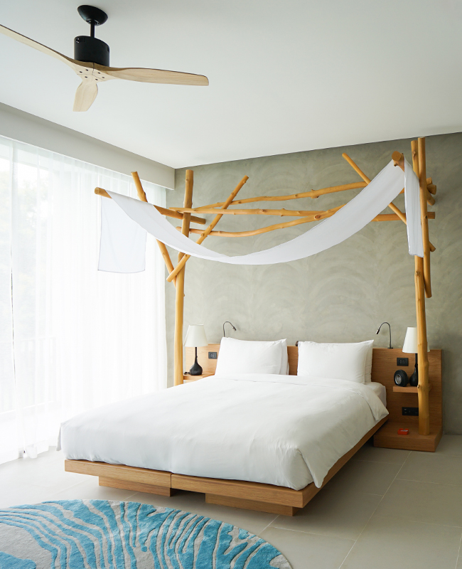 A serene bedroom with a wooden canopy bed, white bedding, sheer drapes, soft lighting, and a modern fan on the ceiling, creating an airy, minimalist atmosphere.