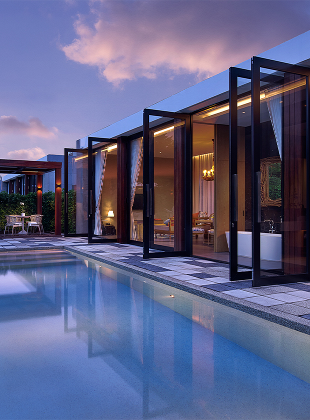Modern house exterior at dusk with open glass doors, a lit interior, pool in the foreground, and greenery surrounding the patio area, creating a serene atmosphere.