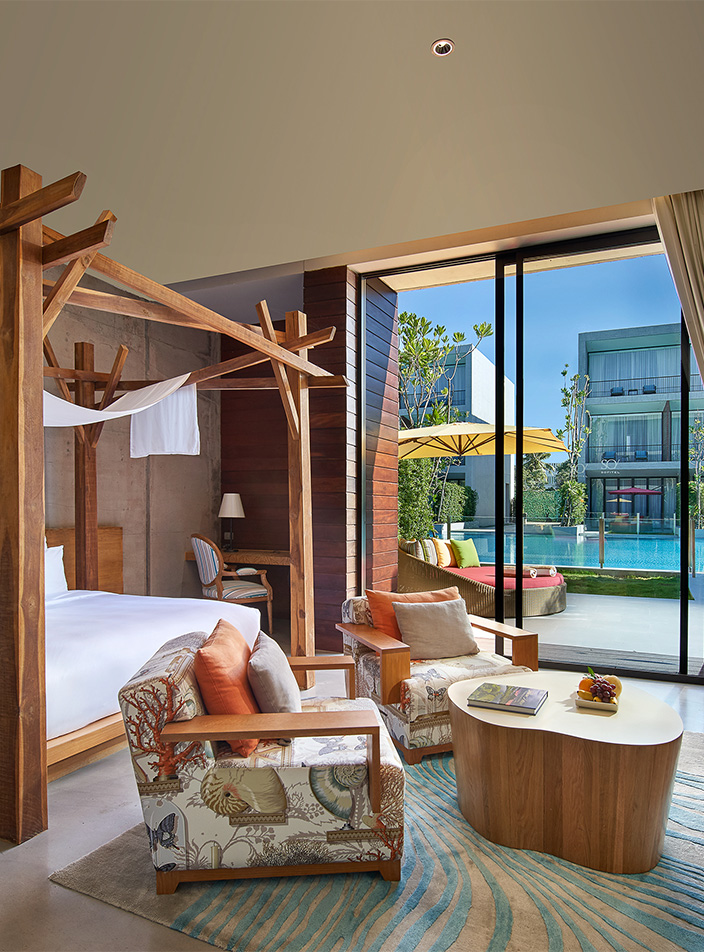 Luxurious bedroom with a large bed, wooden furniture, and cozy armchairs. A glass door opens to a vibrant pool area, creating a serene and inviting atmosphere.