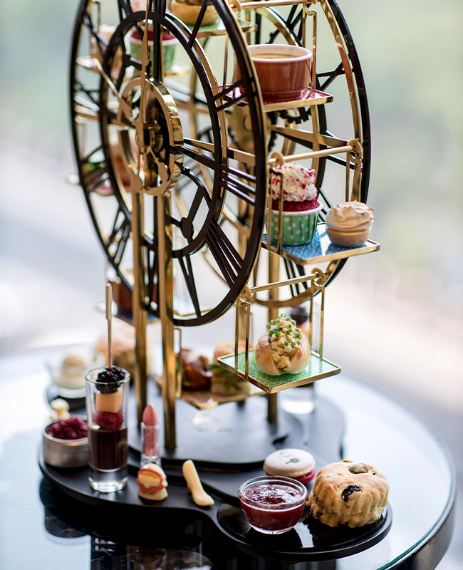 A selction of sandwiches and desserts hang from a serving tray in the shape of a clock. Other cakes surround it on a table.