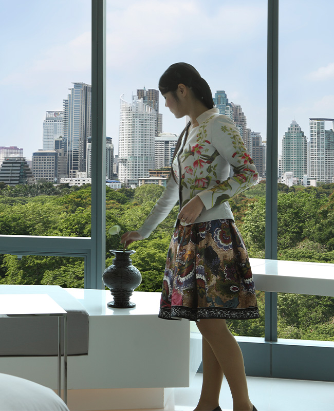 A woman places a flower in a black vase in front of a floor to ceiling window with a beautiful view of Bangkok.
