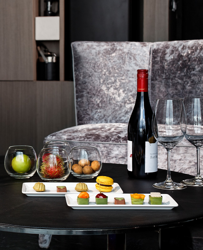 A wine bottle, two glasses, two plates of small food items and four glasses filled with various objects sit on a black coffee table in front of a great couch.