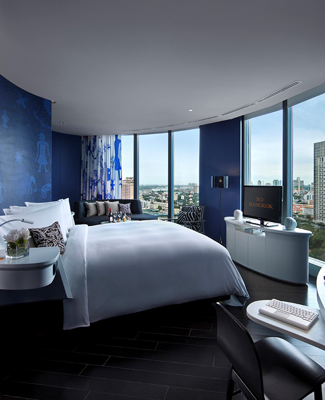 A large blue hotel room with a kind bed facing floor to ceiling windows, a desk, couch and flatscreen TV.