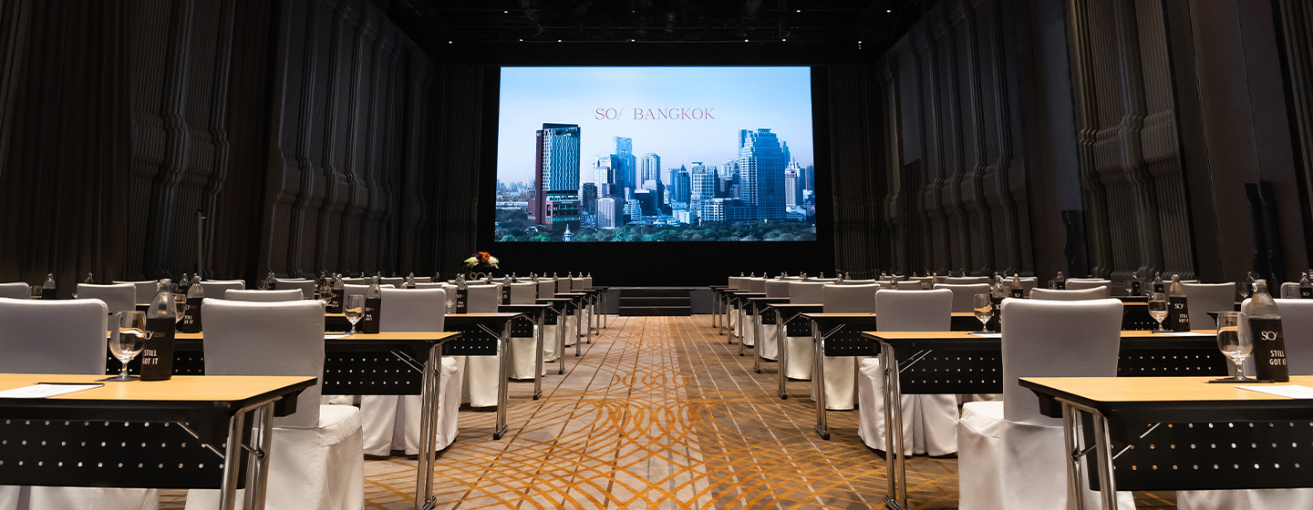Rows of meeting tables and chairs sit in a high-ceilinged event hall facing a large screen.