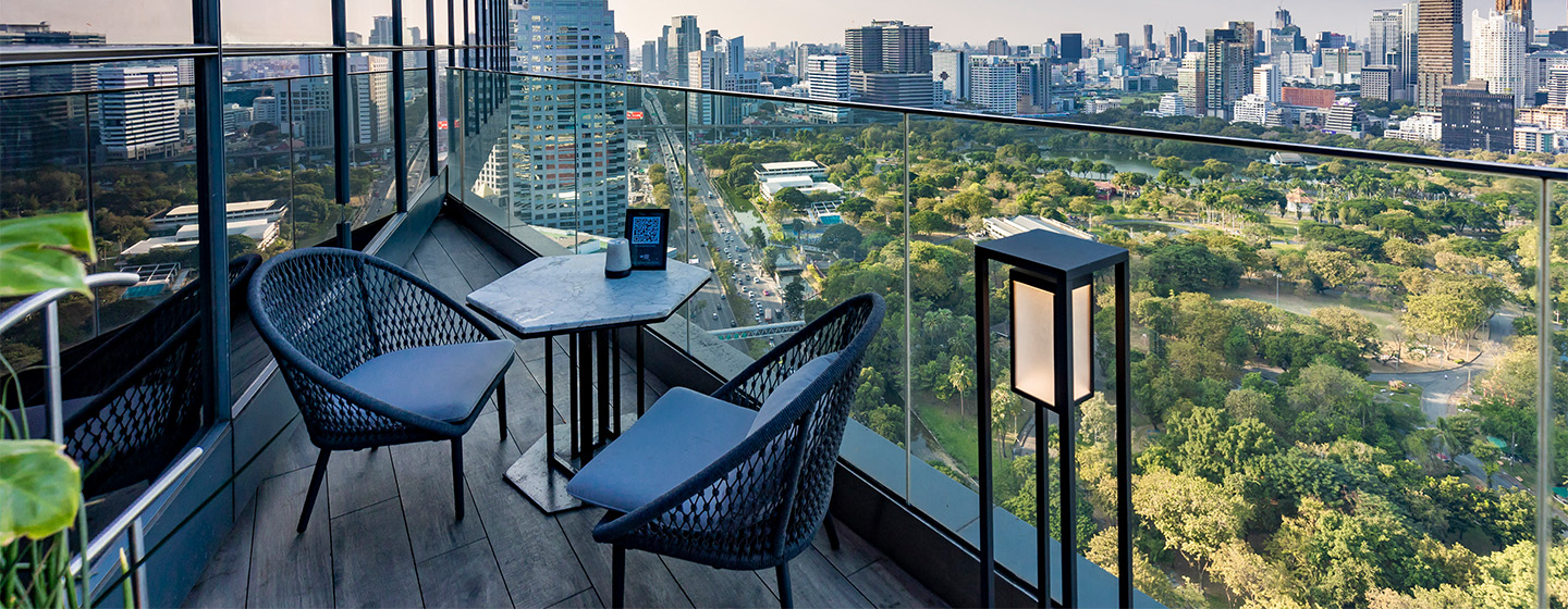 A table and two chairs sit on a balcony overlooking a beautiful view of Lumpini Park and the Bangkok skyline.