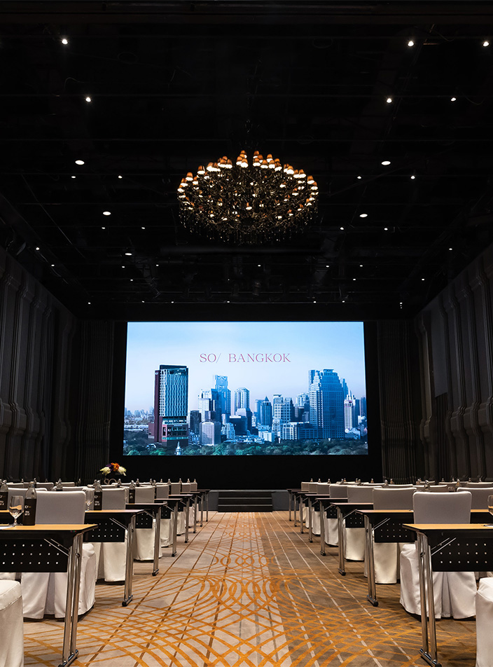Rows of meeting tables and chairs sit in a high-ceilinged event hall facing a large screen.