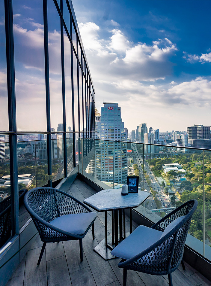 A table and two chairs sit on a balcony overlooking a beautiful view of Lumpini Park and the Bangkok skyline.