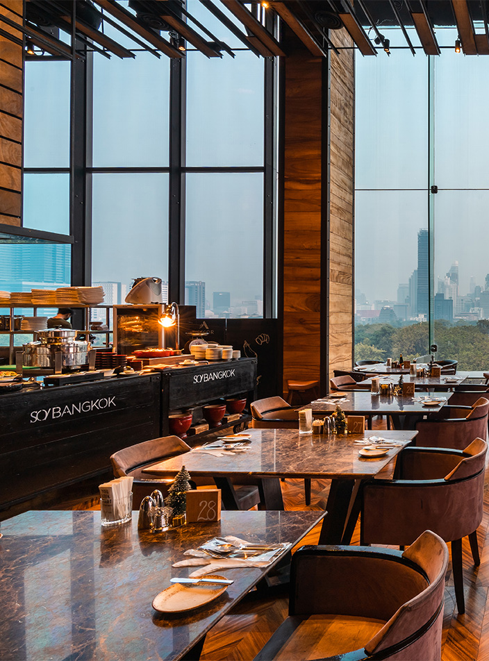 A large restaurant. There are multiple dining tables and chairs and a buffet table and floor-to-ceiling windows overlook Bangkok's Lumpini Park.