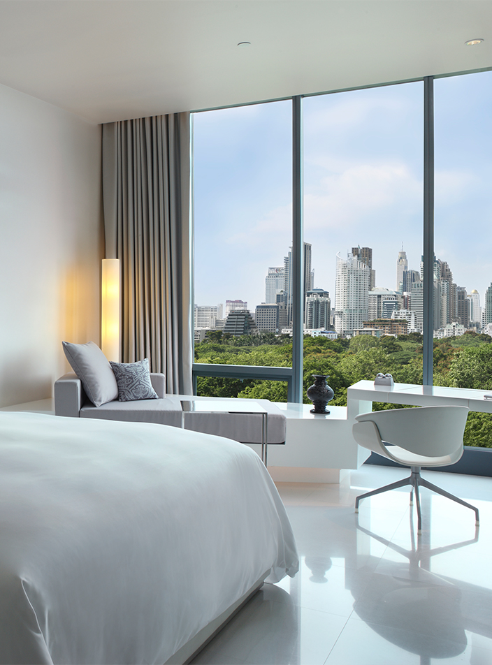 A hotel room with a large bed, desk, chaise longue and floor to ceiling windows with a view of the Bangkok skyline.