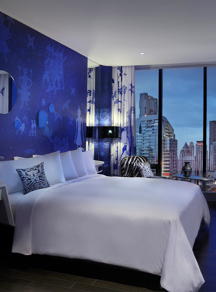 A blue and white themed hotel room with a large bed, desk, and floor to ceiling window displaying a view of Bangkok.