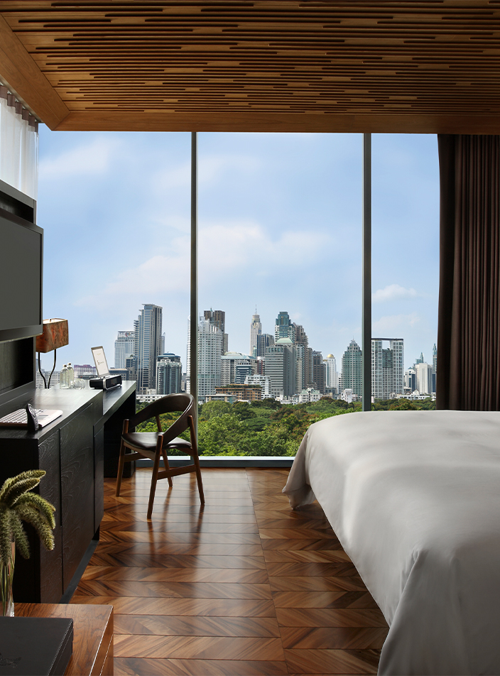 A hotel room with a bed, desk area and large floor-to-ceiling windows overlooking Bangkok's Lumpini Park.