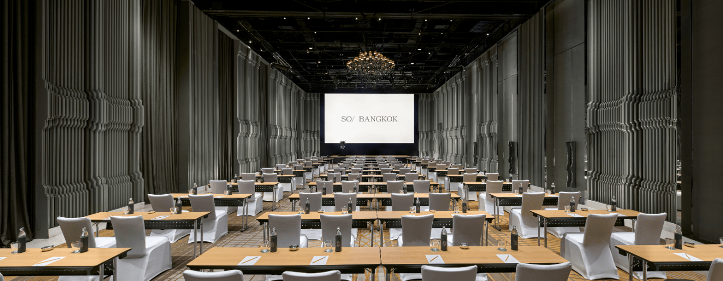 Conference room with rows of tables and white-covered chairs facing a large screen displaying a Bangkok cityscape and the words 'SO/ Bangkok'.